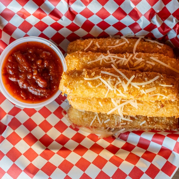 Golden-brown mozzarella sticks topped with shredded cheese, served with a side of marinara sauce in a red-checkered basket.